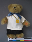 Basset the teddy bear with swivel head and jointed limbs, label at back 'Salco House, River Way, Harlow, Essex, bronze neck ribbon, homemade black gown and white surplice, yellow 'counter' medal with faded 'B', black cap with red decoration.
