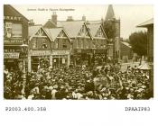 Sepia postcard showing view looking north east from Winton Square, Basingstoke at top of Sarum Hill on the 20th July 1908, a large crowd of men, women and children are surrounding three cars, in the back of the central car is standing, with long white b