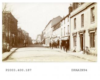 Sepia postcard showing view looking west along London Street, Basingstoke, to the left two women with long coats and large hats walking away on pavement, in front of them Burberrys clothing factory and shop, to the right is a sign for tea and refreshmen
