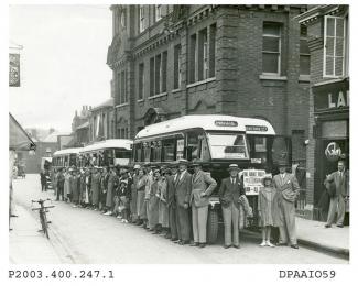 Sepia photograph showing large number of employees from John Mares clothing factory about to set off on a factory outing, view looking north east along New Street, Basingstoke with large group of adults and some children, men are wearing jackets, some w