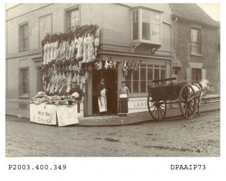 Sepia photograph showing view looking south east at junction of Winchester Street, Basingstoke, on left and Victoria Street on right, on the corner is the 19th century shop and premises of George James Lansley, butcher, the shop has a large display of l